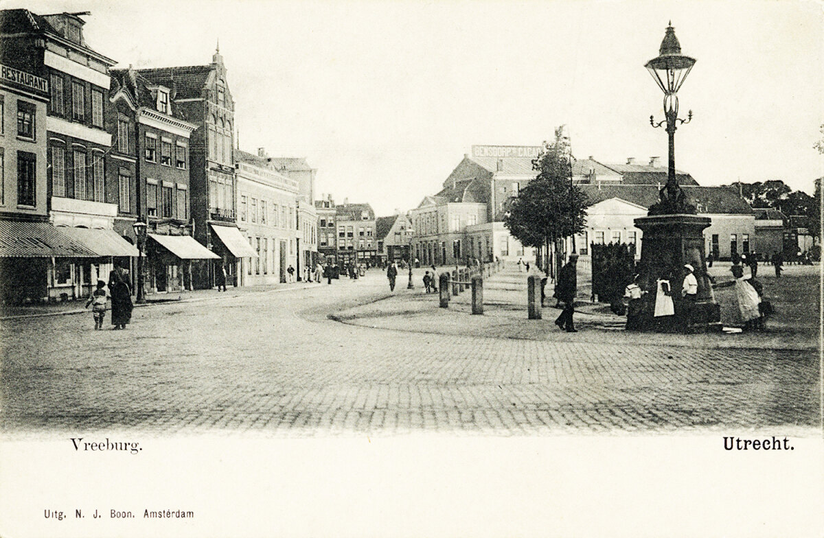 Rond 1900. De fotograaf staat met zijn rug naar waar tegenwoordig de hoek met de St. Jacobsstraat is en we kijken richting het zuiden. In het smalle derde pand links zit de poort naar de Zakkendragershof, destijds de tuin van het oude mannen- en vrouwenhuis Oudaen. Tussen de twee huizen rechts daarnaast zien we de ingang van de Zakkendragerssteeg. Rechts op de voorgrond zien we de grote pomp op de hoek van het plein met een grote gaslantaarn erop. De pomp was toen al aangesloten op de waterleiding. Links erachter zien we een urinoir, maar ik plaats de foto hier natuurlijk omdat we op de achtergrond de schouwburg in zijn geheel kunnen zien staan en goed kunnen zien wat een imposant gebouw het toen al was. HUA2683