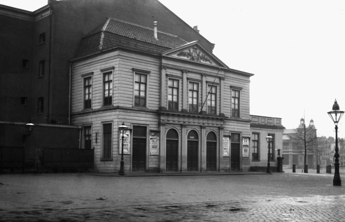 De schouwburg in 1905. De foto is genomen vanaf het punt waar het Vredenburg overgaat in de Lange Elisabethstraat. Rechts ervan zien we nog een stukje van de Fruithal. We zien hier de schouwburg zoals deze ontstaan is na de verbouwing in 1880/81. Hier is goed zichtbaar dat het front van het oude houten gebouw uit 1820 is blijven staan en er een grote hal achter tegenaan is gebouwd.