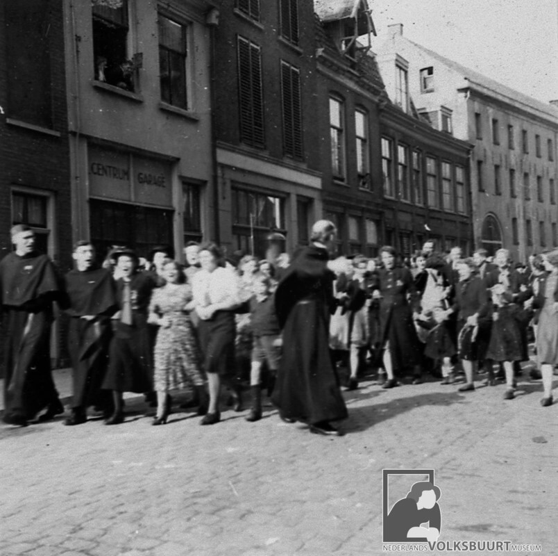 Pastoor Van Nuenen kijkt even achterom of iedereen in de Waterstraat nog meeloopt in een feestelijke optocht om de komende bevrijding te vieren. Foto: VBM