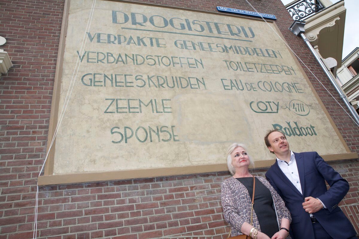 Wethouder Kreijkamp en bewoner Sylvia Six bij de herstelde muurreclame. Foto: Ton van den Berg